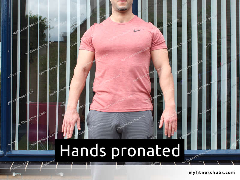 A fit man wearing workout clothing while standing outside in front of a window, with his hands pronated.