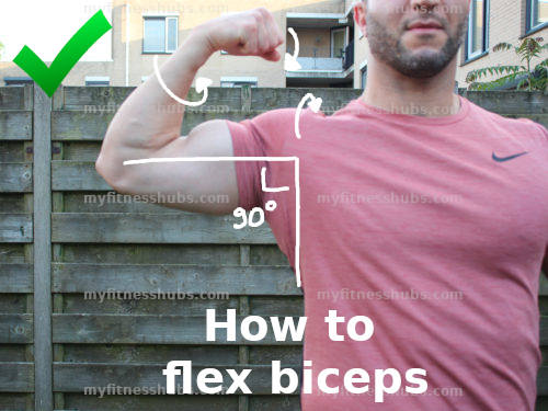 A close up of a man in a fitness T-shirt doing a biceps flex while standing outside in what looks to be a garden.