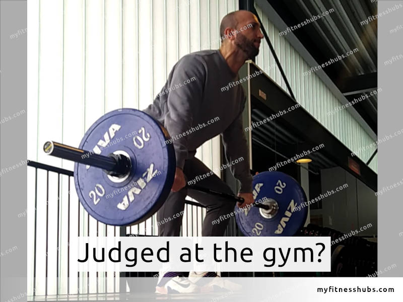 A front-side view of fit man deadlifting in a gym on a weightlifting platform, with the text overlay 'judged at gym?'