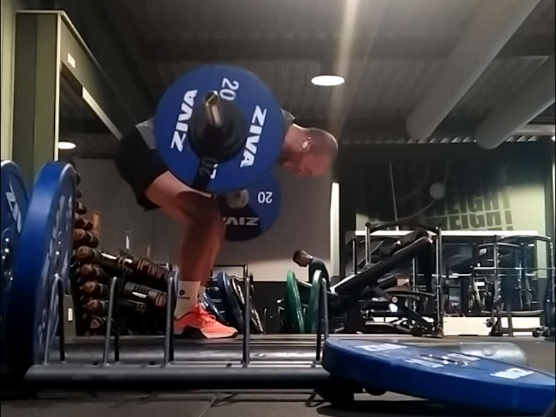 A man doing a heavy Pendlay row with a barbell loaded with weights while standing on a weightlifting platform in a commercial gym.