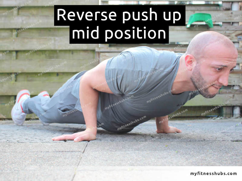 A view of a front angle view of an athletic man in the middle position of a reverse push up done outdoors.