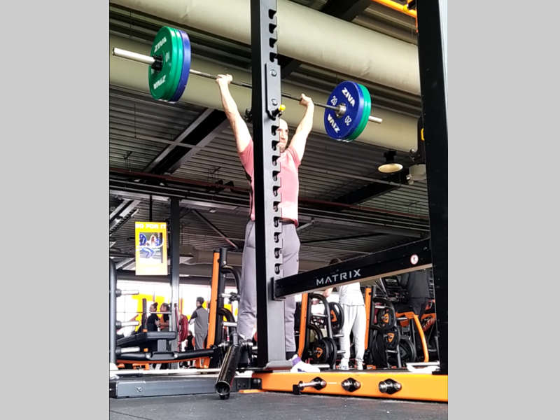 A young man doing a standing barbell overhead press in front of a squat rack in the gym.