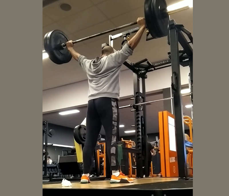 A man in a gym doing a vertical push exercises by holding a barbell overhead with wide grip.